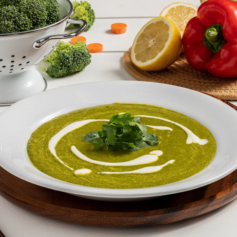 Green soup with cilantro and a cream swirl in a white bowl, with lemon, broccoli and red pepper in the background.