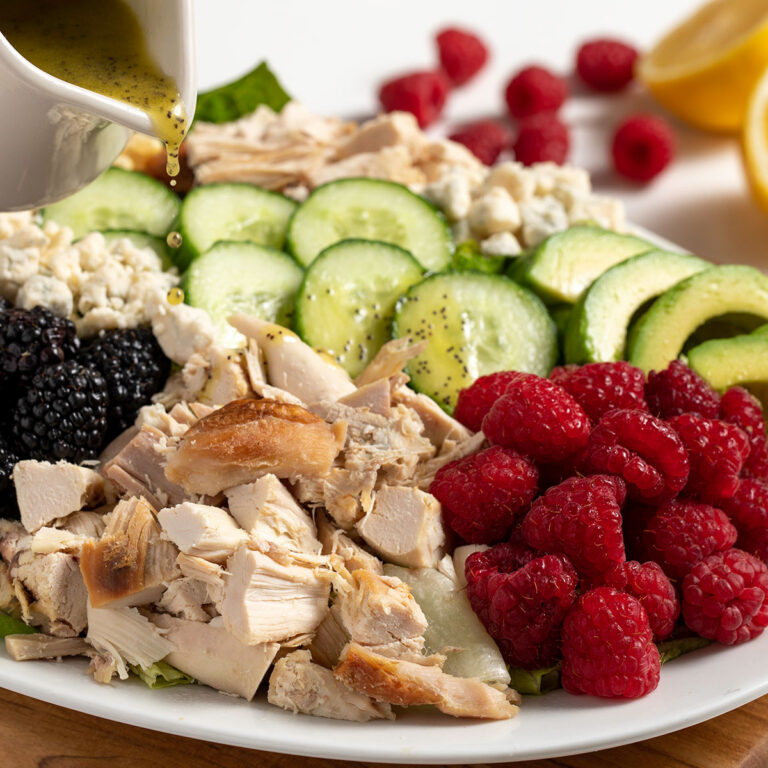 Plate of salad with shredded chicken, cucumber rounds, raspberries, blackberries, avocado, and feta, as a dressing is poured over it.