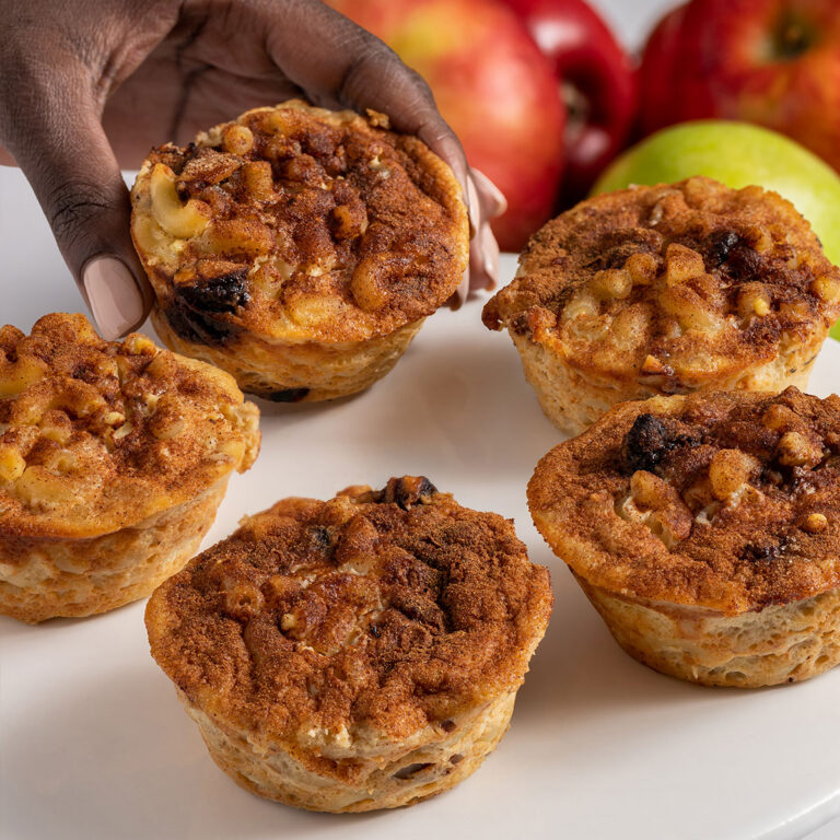 Hand reaching for a cinnamon apple muffin with crumb topping on a white plate, apples blurred in the background