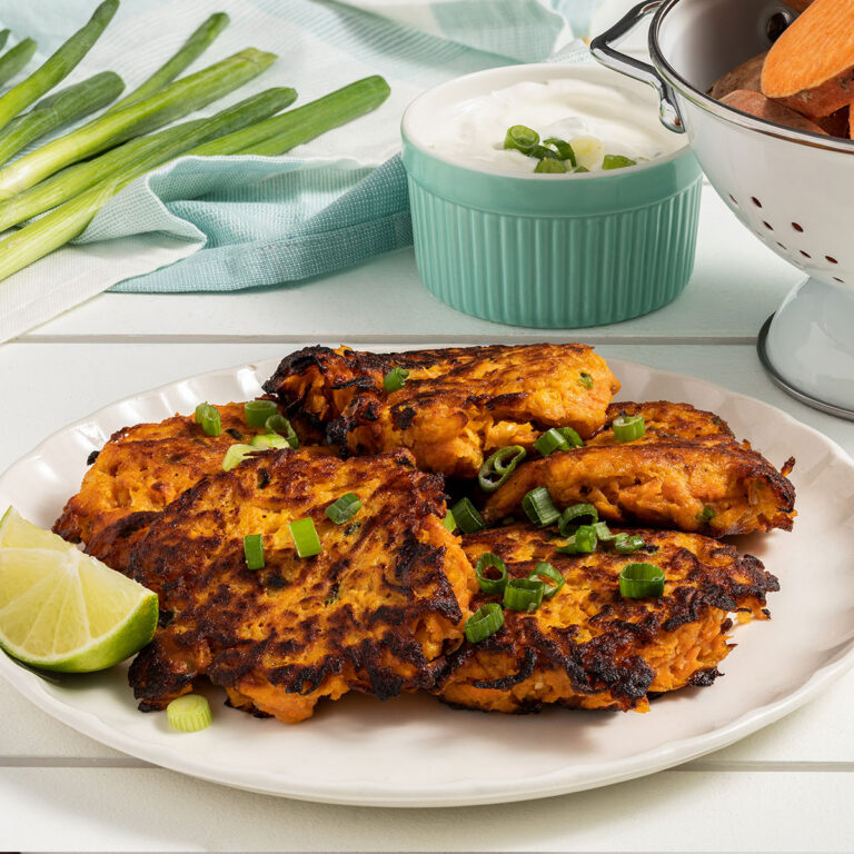 Crispy browned salmon patties garnished with green onions on a white plate, lime wedges nearby, with dip and asparagus in the background