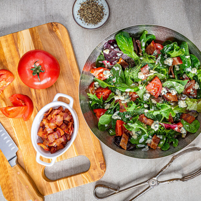 Large metal bowl filled with mixed greens, tomatoes, croutons, and feta, with a cutting board showing chopped tomatoes and a white dish of bacon bits nearby.
