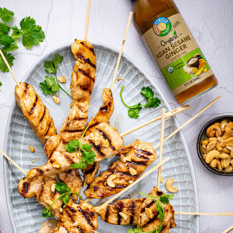 Skewered grilled chicken pieces with cilantro, sitting on a platter beside a bottle of organic Asian sesame ginger dressing and a small bowl of cashews.