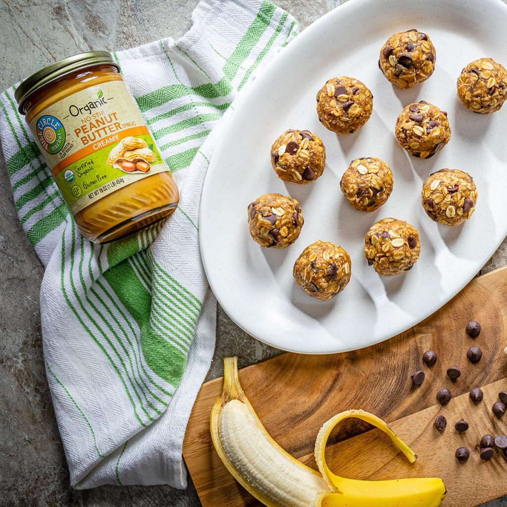 Jar of organic peanut butter next to a plate of oat-and-chocolate energy bites on a white platter with a banana nearby.
