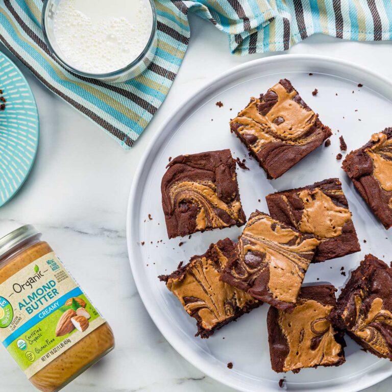 Brownie squares swirled with peanut butter on a white plate, nearby almond butter jar and a glass of milk on a marble surface.